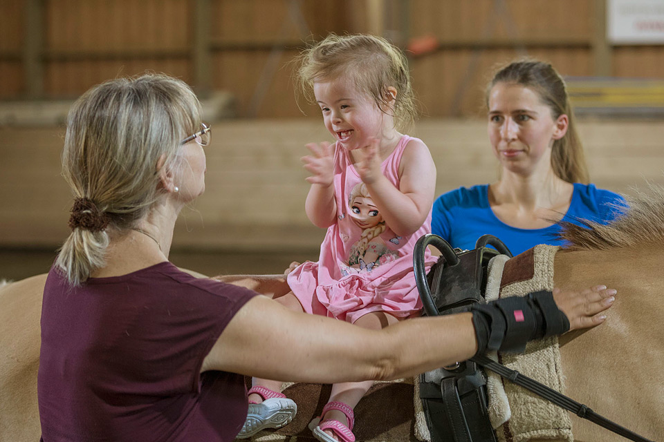 Ein Kind sitzt auf einem Therapiepferd. Die Therapeutin spricht mit dem Kind, während die Mutter im Hintergrund zusieht.